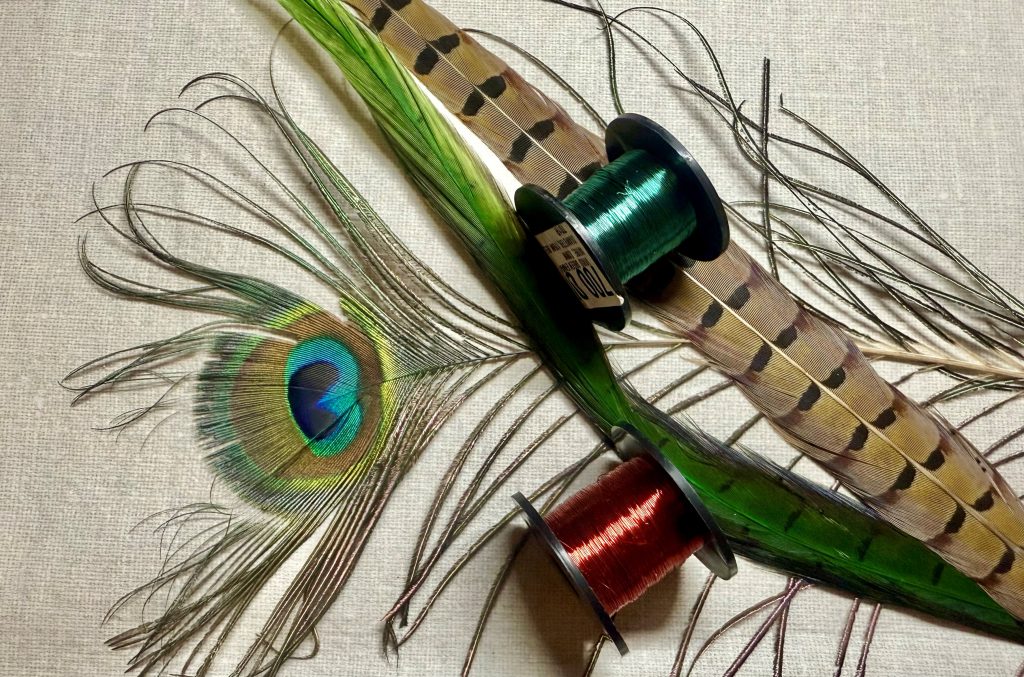 Material for tying Pheasant tail nymph flies arranged on a tabletop including peacock feathers and spools of fine coloured copper wire